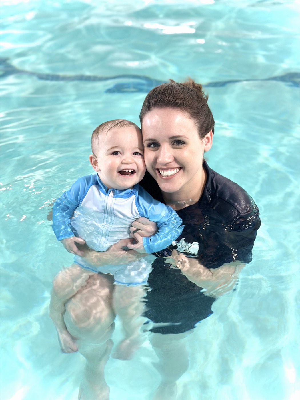 Instructor Katrina smiling in the pool while holding a happy baby in a swim shirt